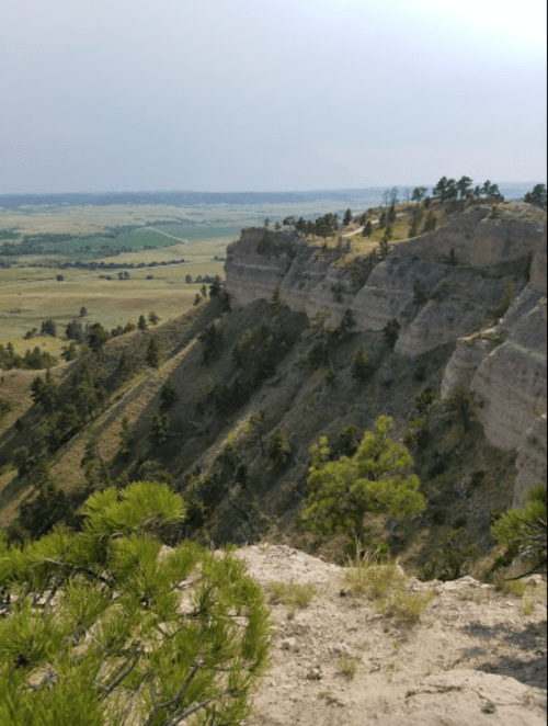 A scenic view of layered cliffs and green valleys under a cloudy sky. Pine trees dot the landscape.