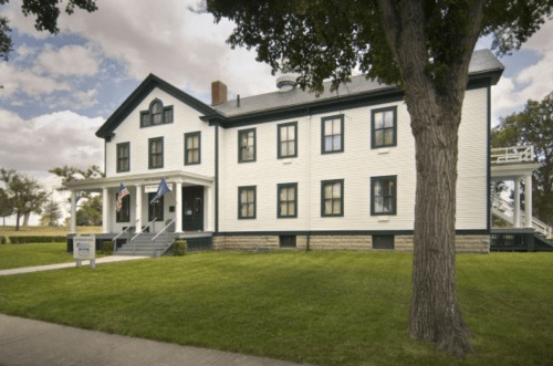 A large, two-story white building with green trim, surrounded by grass and trees, featuring flags at the entrance.