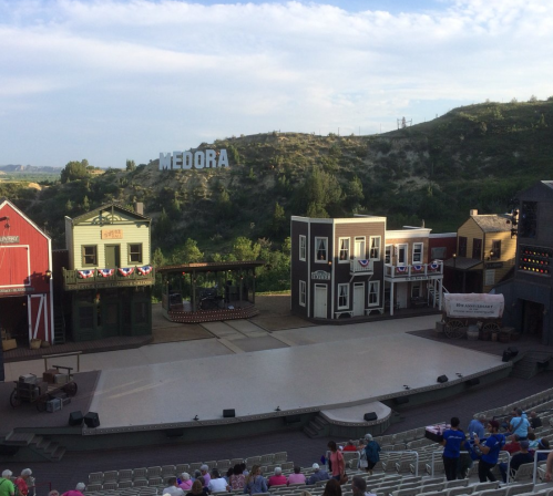 A stage set with Western-style buildings, empty seats, and a hillside sign reading "MEDORA" in the background.