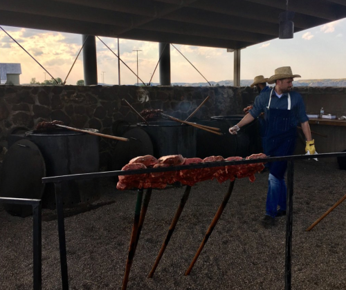 A cook in a straw hat tends to meat roasting over an open fire under a shaded structure.