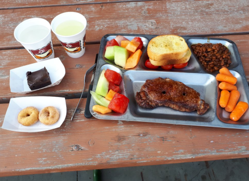 A metal tray with steak, toast, baked beans, fruit, carrots, and two cups of drink, alongside dessert items.