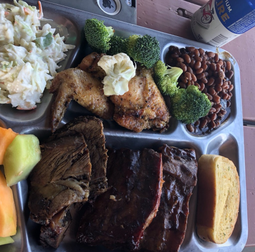 A metal tray with grilled chicken wings, broccoli, baked beans, sliced meats, cornbread, and a side of coleslaw and melon.