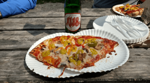 A slice of pizza topped with peppers and mushrooms, next to a green bottle on a picnic table.