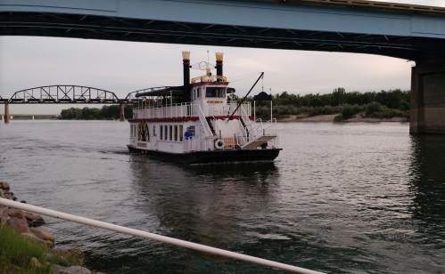 A paddleboat navigates a river beneath a bridge, with trees lining the shore in the background.