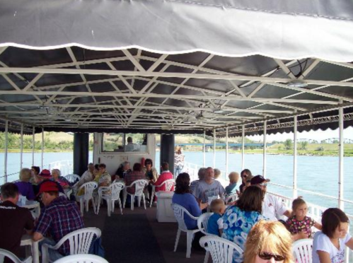 A crowded boat interior with people sitting at tables, enjoying a scenic view of the water and shoreline.