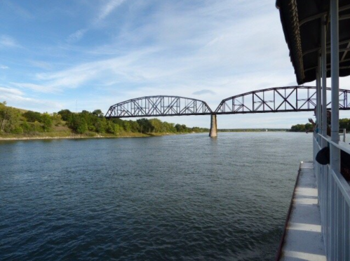 A river flows beneath a metal bridge, surrounded by greenery and a clear blue sky.