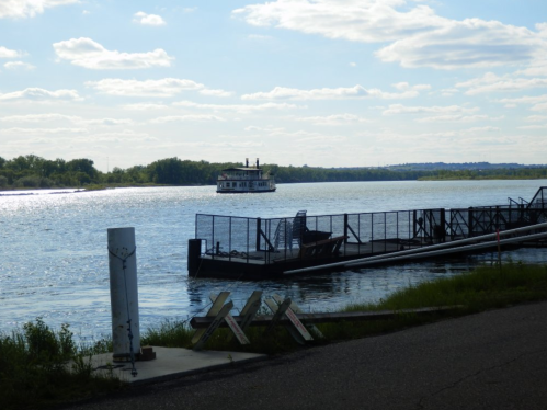 A river scene with a boat in the distance, a dock in the foreground, and a clear blue sky with scattered clouds.