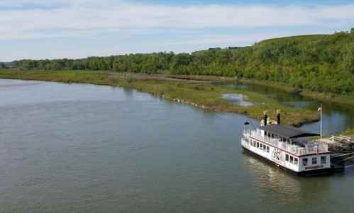 A riverboat docked by a lush green riverbank under a clear blue sky.