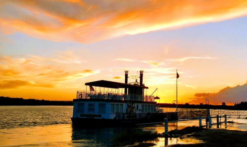 A riverboat docked by the shore at sunset, with vibrant orange and purple clouds reflecting on the water.
