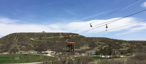 A scenic view of a hill with a zip line, surrounded by greenery and a clear blue sky.