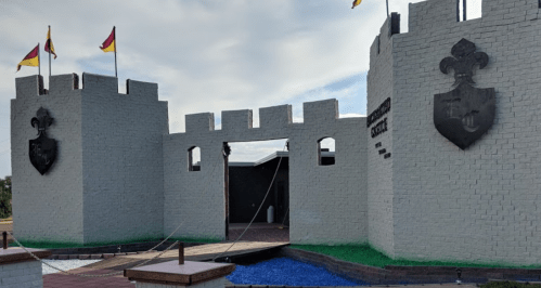 A large, white castle-like structure with flags and shields, featuring a bridge over water leading to an entrance.