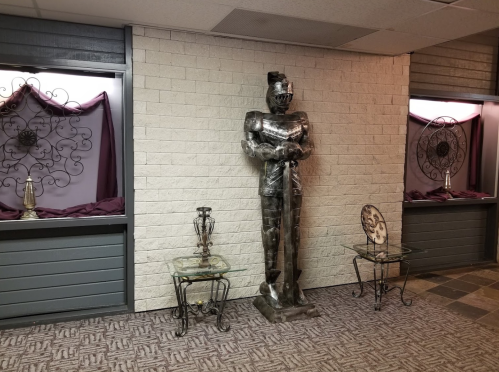 A metallic statue stands in a hallway, flanked by decorative tables and windows with purple drapes.
