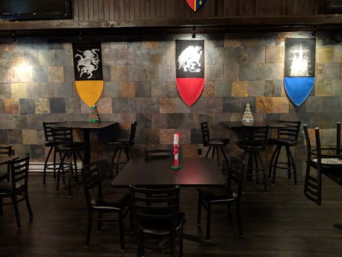 A dimly lit restaurant with black tables and chairs, featuring colorful shields on a stone wall.