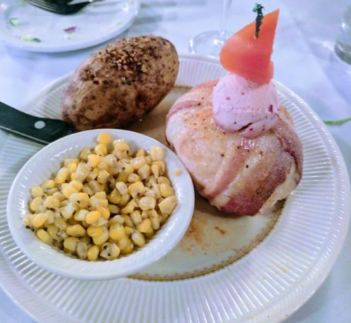 A plate with a bacon-wrapped meatball topped with a tomato, served with corn and a baked potato.