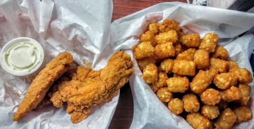 A plate with crispy fried chicken strips and a side of golden tater tots, served in paper wrappers.