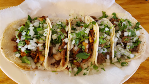 A plate of four tacos filled with meat, topped with chopped onions and cilantro, served on a white paper.