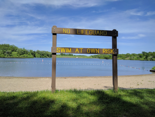Sign by a sandy beach near a lake, warning "No Lifeguard" and "Swim at Own Risk" under a clear blue sky.