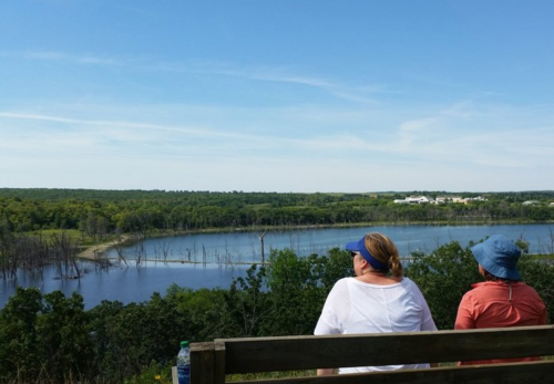 Two people sit on a bench overlooking a serene lake surrounded by greenery under a clear blue sky.