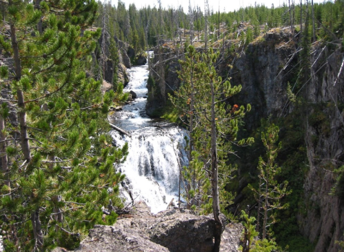A cascading waterfall flows through a rocky canyon, surrounded by tall green trees and rugged terrain.
