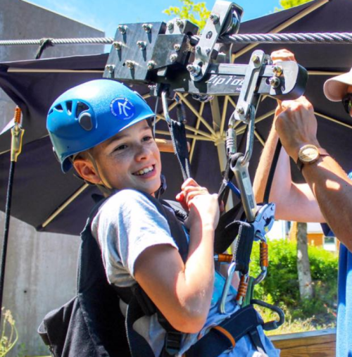A young boy in a blue helmet smiles while preparing for an outdoor zipline adventure.