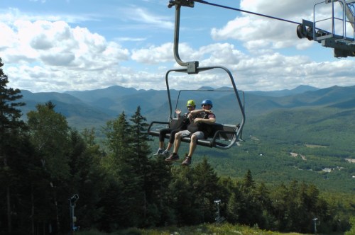 Two people sitting on a ski lift, surrounded by lush green mountains and a blue sky with fluffy clouds.