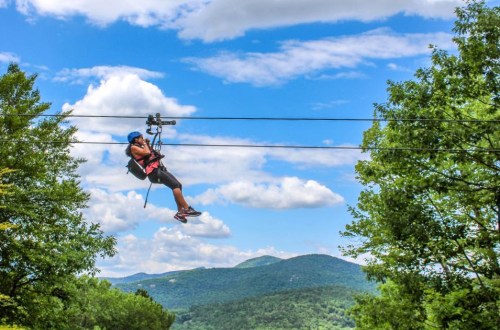 A person zip-lining across a scenic landscape with mountains and blue skies in the background.