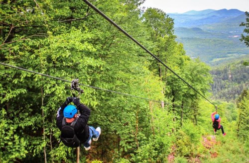 Two people ziplining through a lush green forest with mountains in the background.
