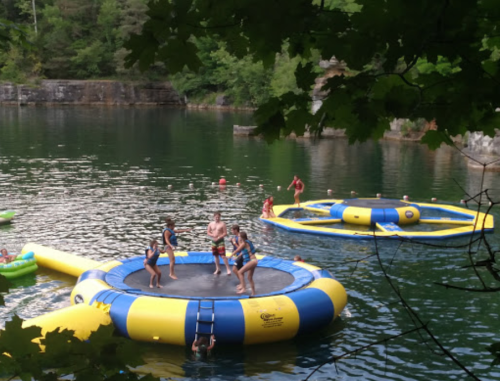 Children and adults enjoy a water trampoline on a lake, surrounded by trees and other inflatable toys.