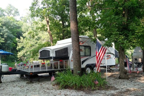 A camper parked among trees, with an American flag and small flags on the ground, in a serene outdoor setting.