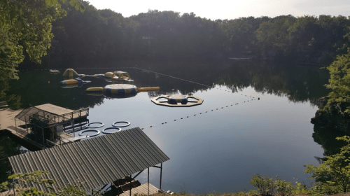 A serene lake with inflatable water toys, surrounded by trees and a wooden dock. The water is calm and reflective.