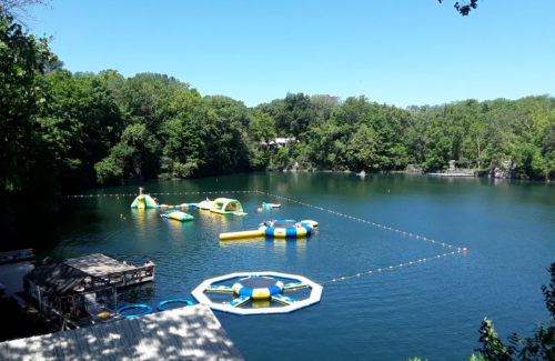 A serene lake with inflatable water attractions and a wooden dock, surrounded by lush green trees under a clear blue sky.