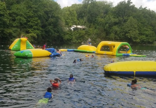 Children playing on inflatable water structures in a lake, surrounded by trees and enjoying a sunny day.