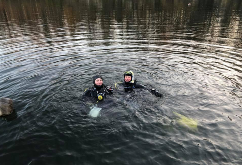 Two divers in wetsuits are floating in calm water, surrounded by ripples and rocky edges.