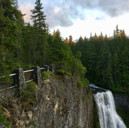 A scenic view of a waterfall cascading over rocky cliffs, surrounded by lush green trees and a cloudy sky.
