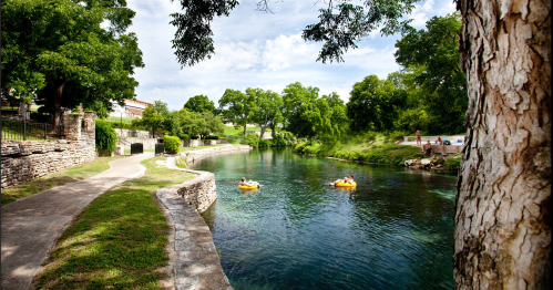 Two people float on inner tubes in a serene river, surrounded by lush greenery and a clear blue sky.