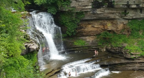 A scenic waterfall cascades over rocky cliffs, surrounded by lush greenery, with people enjoying the water below.