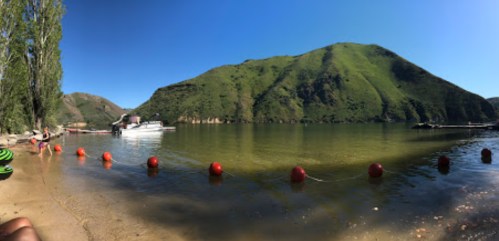 A serene lake surrounded by green hills, with a sandy shore and red buoys marking a swimming area.
