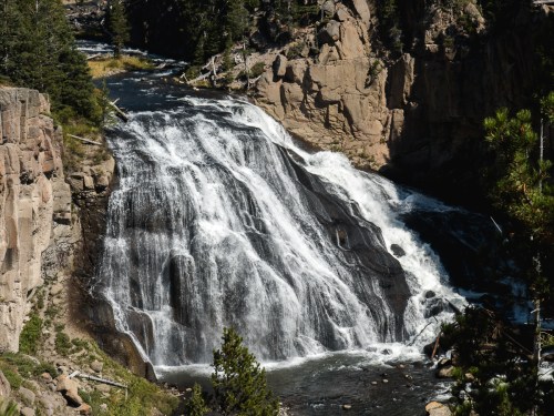 A cascading waterfall flows over rocky cliffs, surrounded by lush greenery and tall trees in a natural landscape.