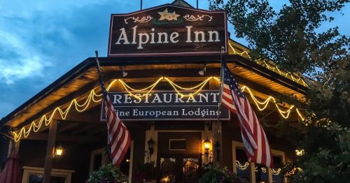The Alpine Inn, a cozy restaurant with a wooden sign and American flags, illuminated at dusk.