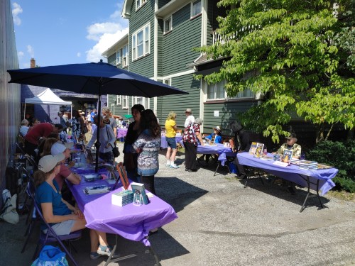 A vibrant outdoor book fair with people browsing tables under umbrellas, surrounded by greenery and buildings.
