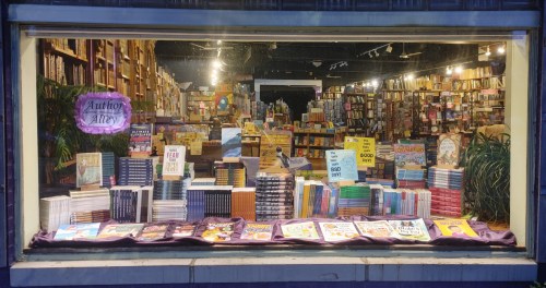 A cozy bookstore window display featuring a variety of books and a sign for "Author Alley" in the background.
