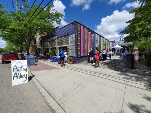 A vibrant street scene with people gathered outside a colorful building, featuring a sign for "Author Alley."