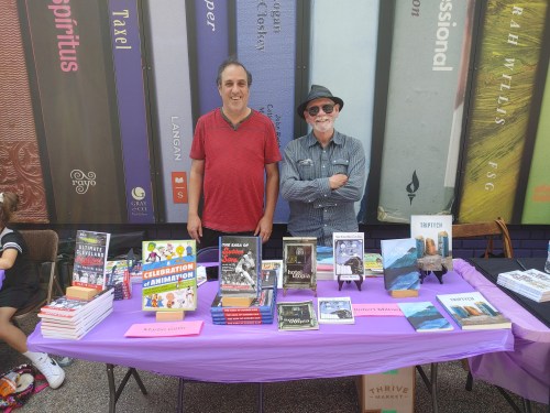 Two men stand behind a table filled with books at an outdoor event, with colorful book spines in the background.