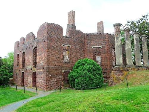Ruins of a brick building with columns, surrounded by greenery and a pathway, under a cloudy sky.