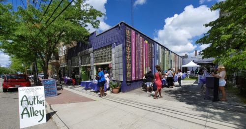 A vibrant street scene with people browsing stalls at Author Alley, surrounded by colorful buildings and greenery.