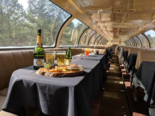 A dining car with tables set for a meal, featuring drinks and a platter of food, surrounded by scenic views.