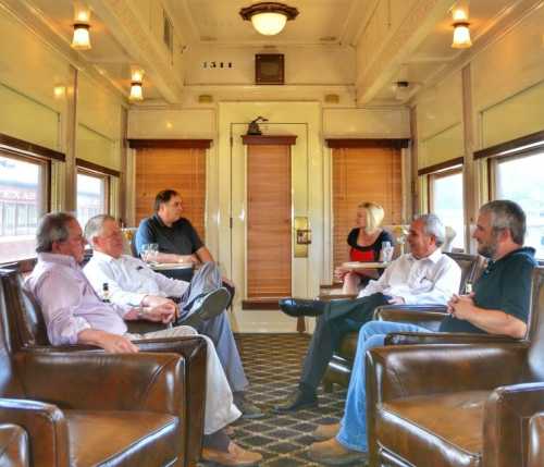 A group of six people seated in a vintage train car, engaged in conversation, with elegant decor and warm lighting.