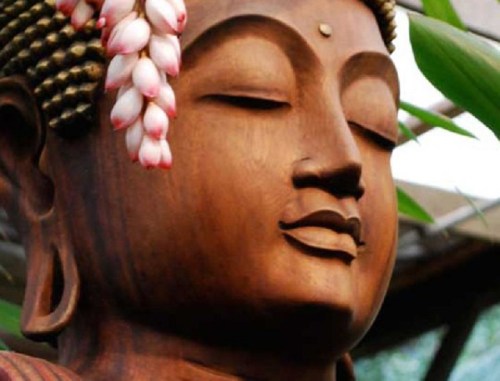 A close-up of a serene wooden Buddha statue adorned with pink flowers, surrounded by green foliage.