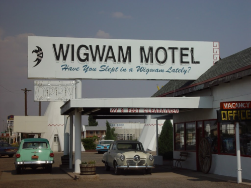 Sign for Wigwam Motel with vintage cars parked outside, featuring a welcoming "Open" message and a retro aesthetic.