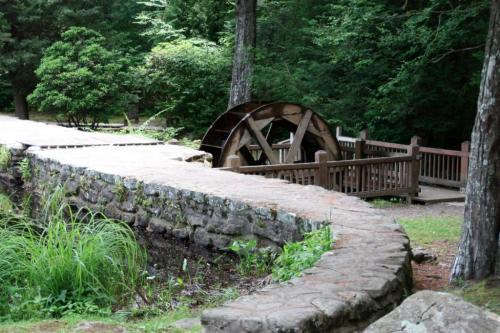 A stone path leads to a wooden water wheel surrounded by greenery in a serene park setting.